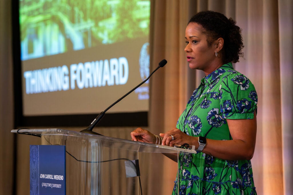 a woman in green stands at a podium in front of a lecture slide titled "thinking forward"